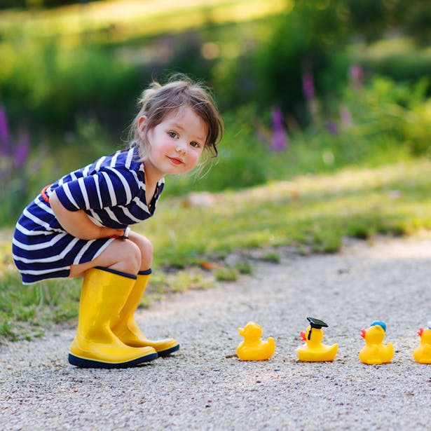 Une petite fille en robe à rayures et bottes de pluie jaunes s'accroupit pour jouer avec une rangée de canards en caoutchouc sur un chemin en gravier, avec une végétation luxuriante en arrière-plan.