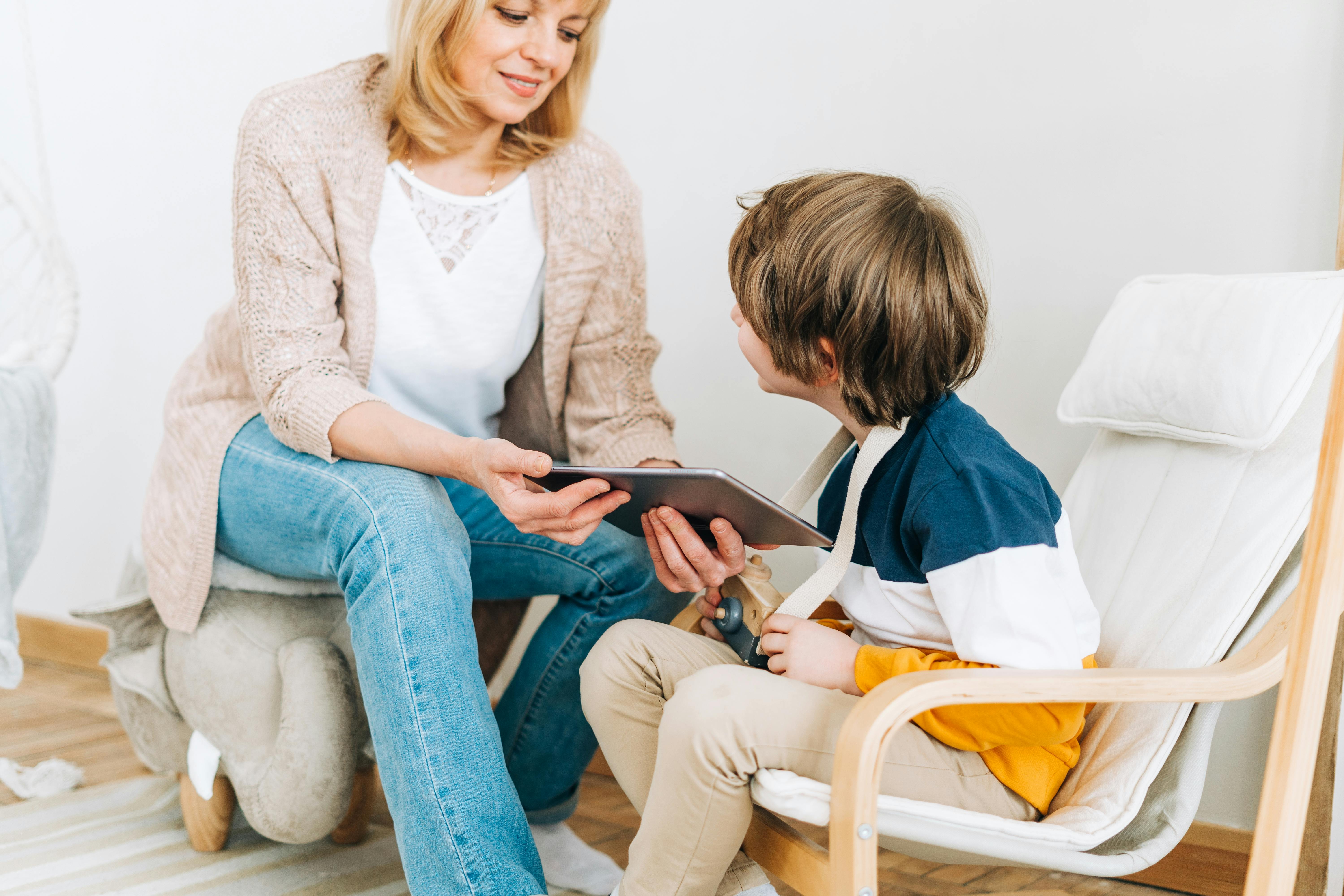 Une mère montre quelque chose sur une tablette à son fils assis sur une chaise.