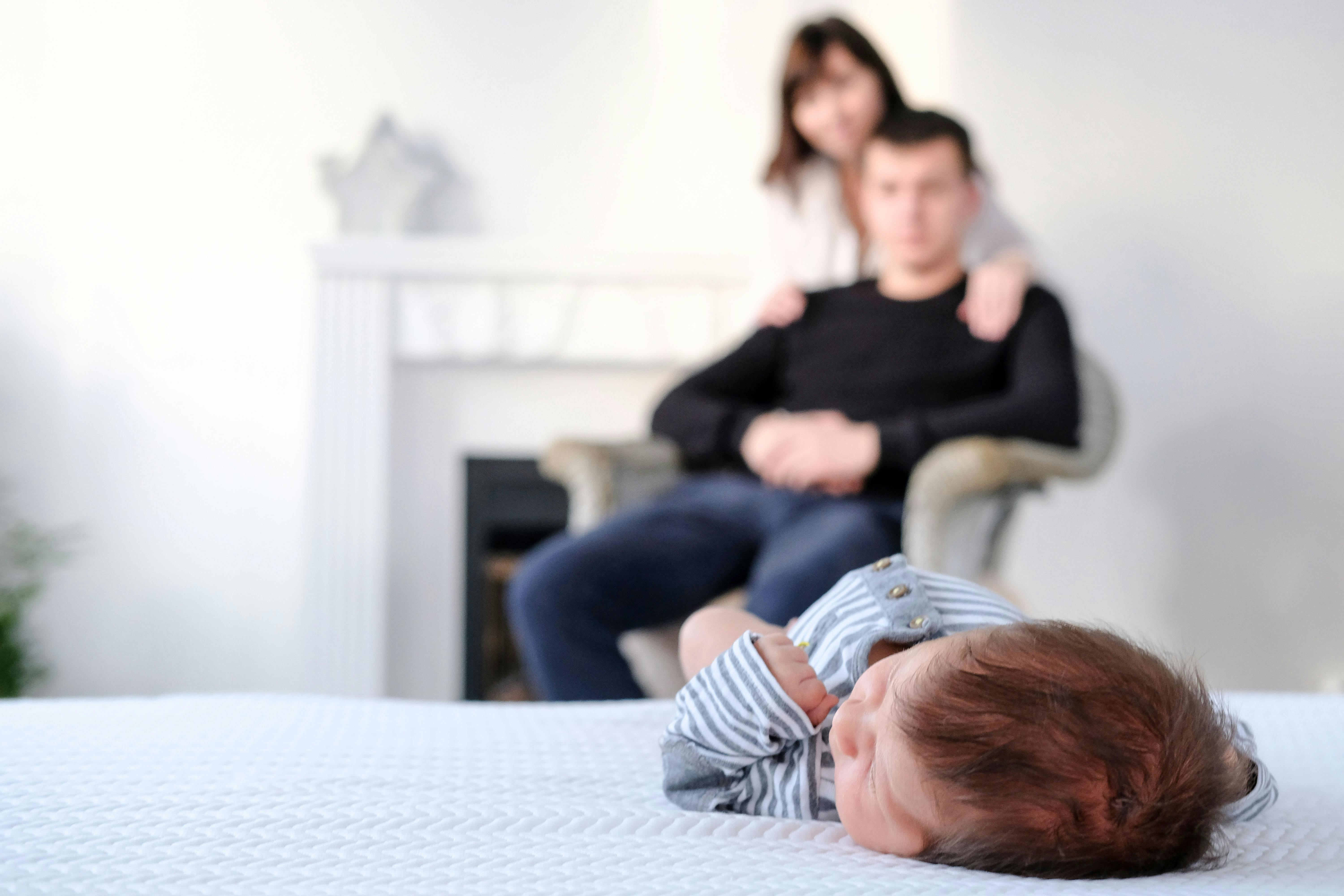 Un bébé est allongé sur un lit avec ses parents en arrière-plan flou.