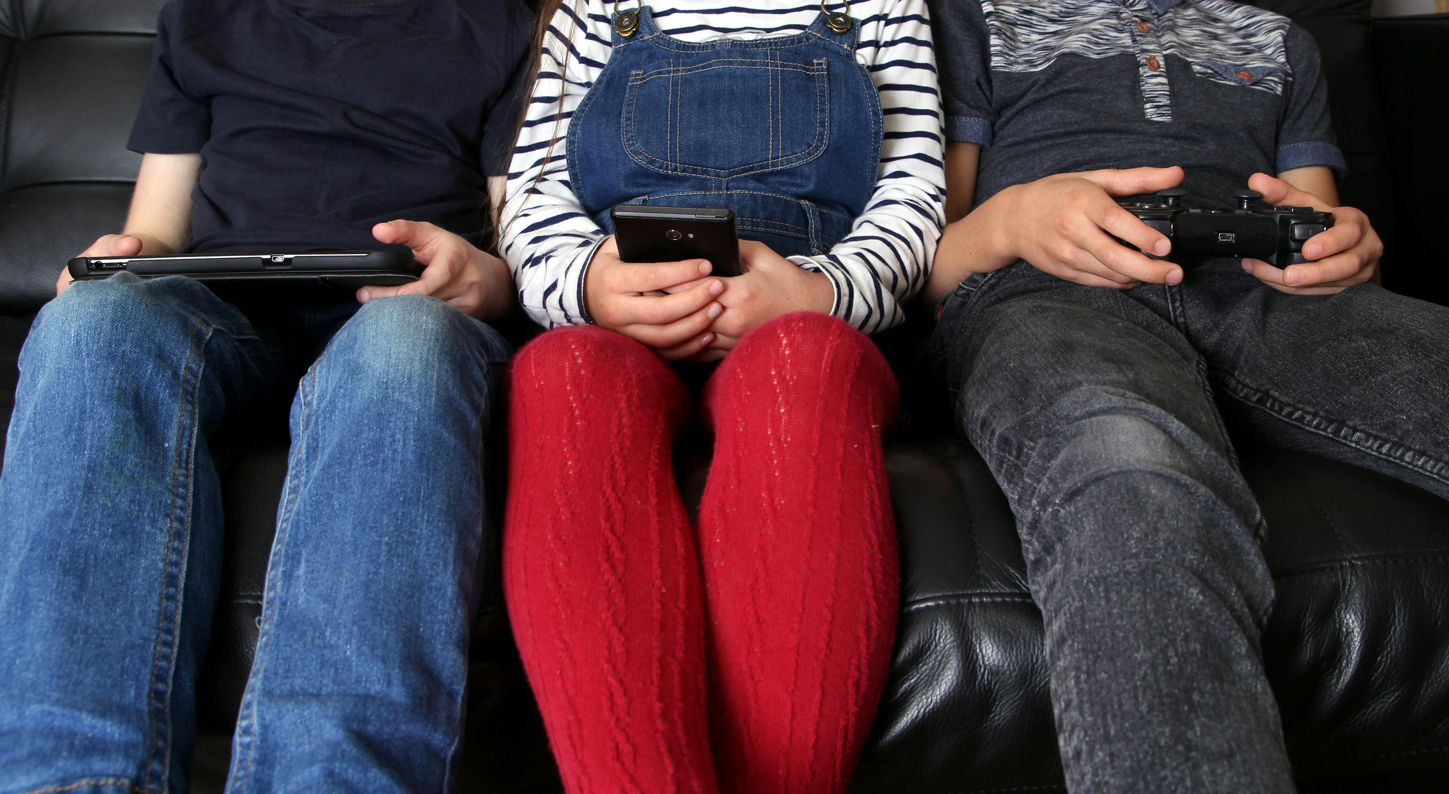 Trois enfants sont assis côte à côte sur un canapé, utilisant chacun un appareil électronique, absorbés par leurs écrans.