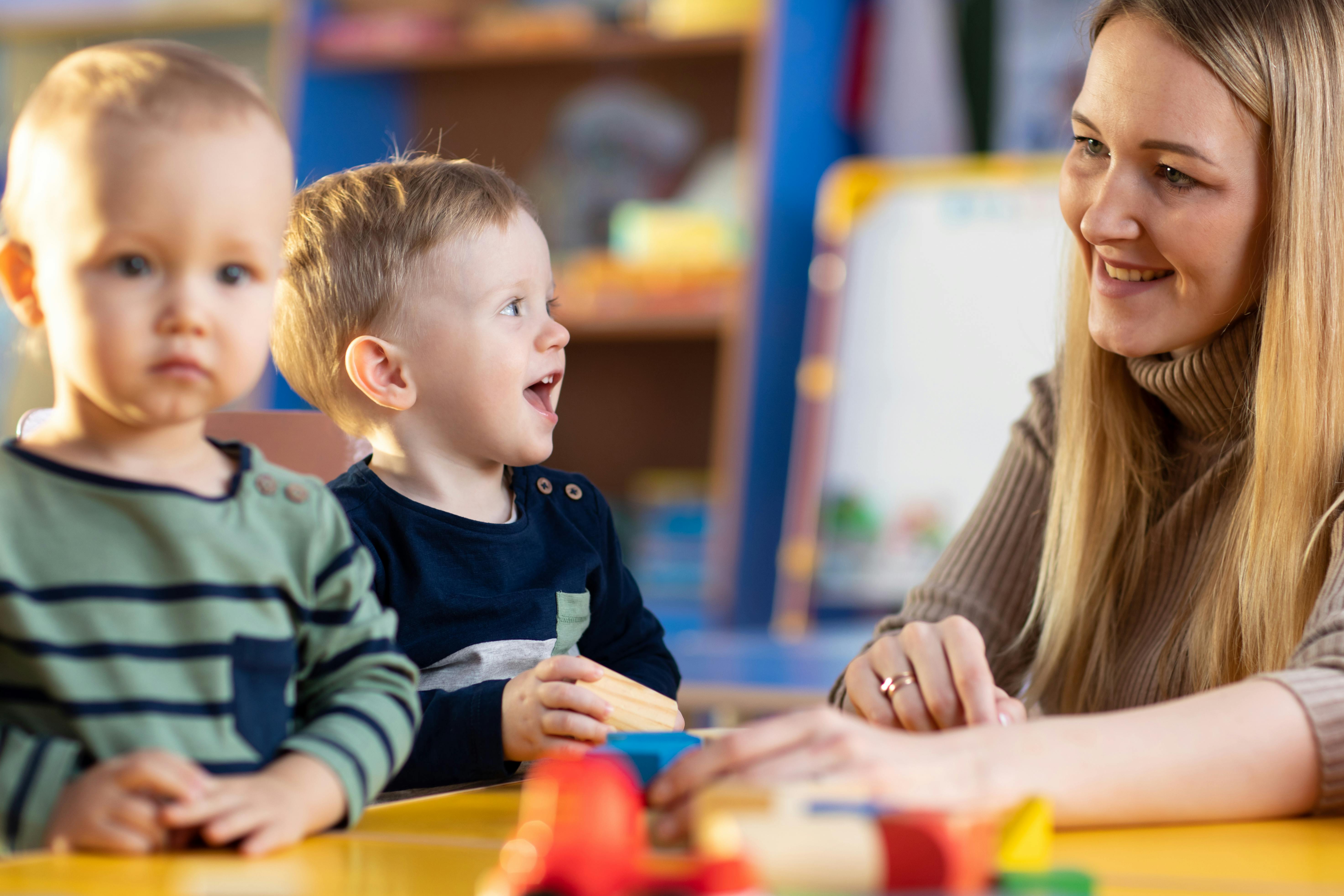 Une enseignante souriante interagissant avec deux jeunes enfants, l'un d'eux riant joyeusement.