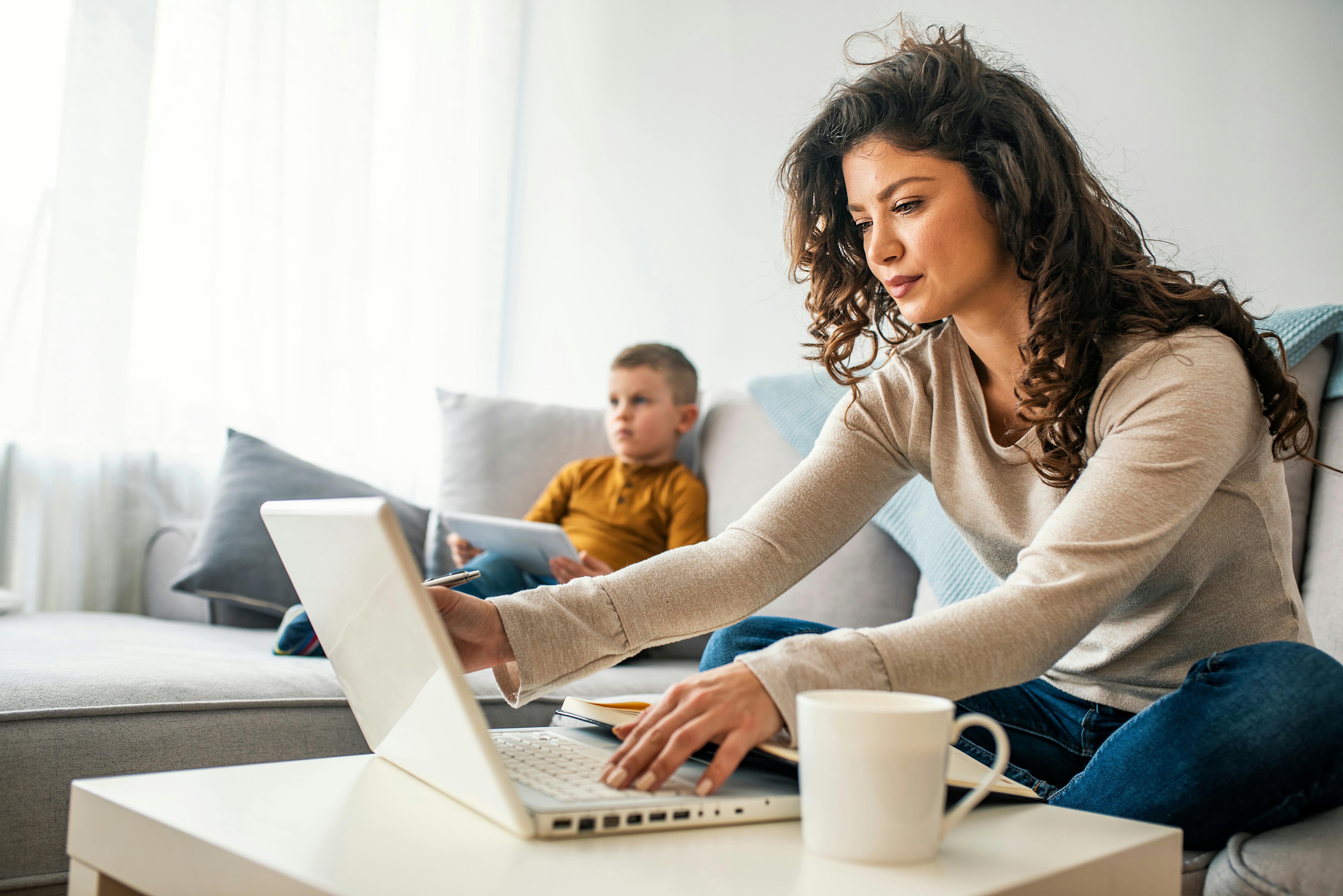 Une mère travaille sur son ordinateur portable pendant que son fils joue sur une tablette à côté d'elle.