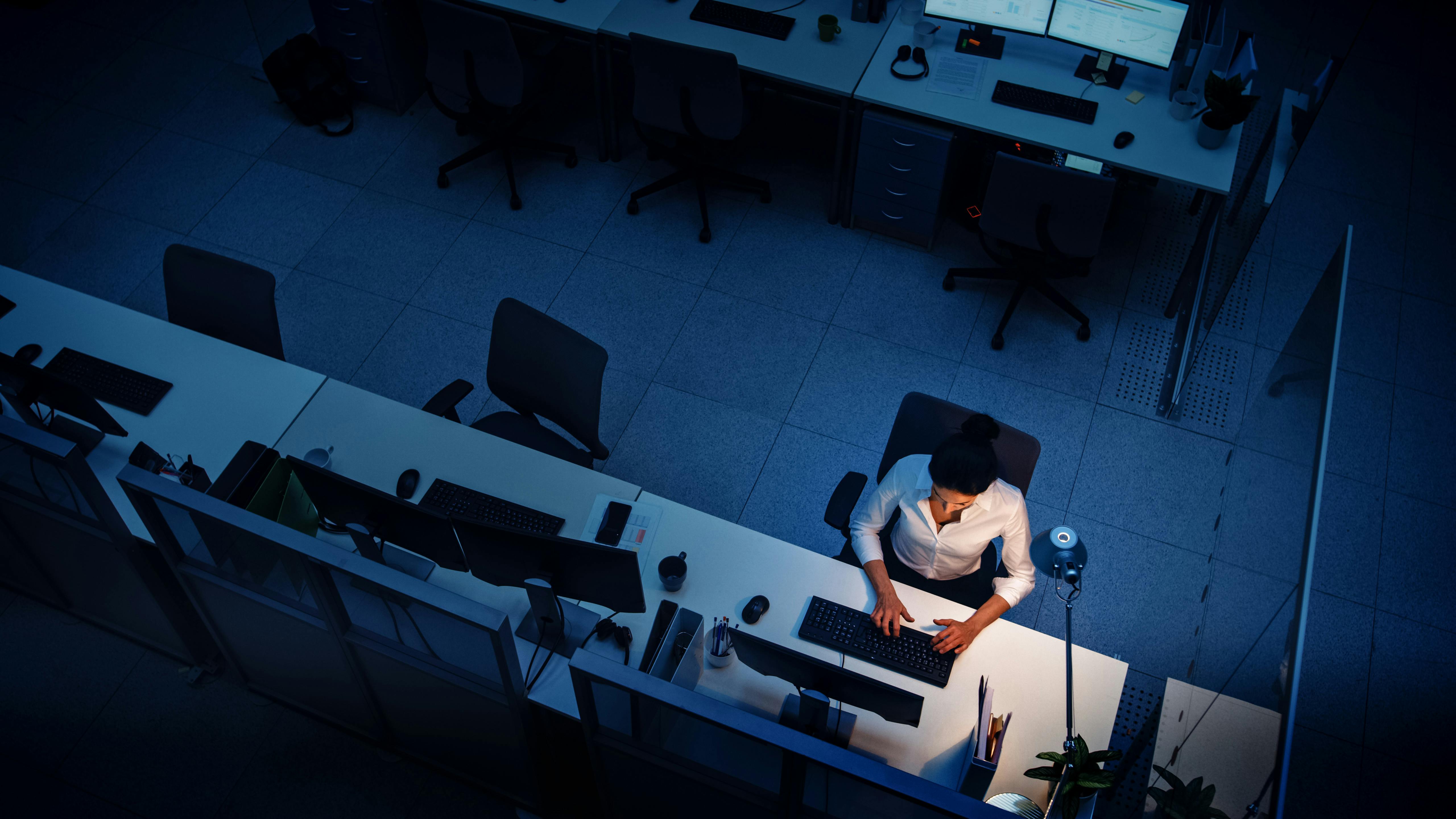 Vue aérienne d'une femme travaillant seule dans un grand bureau éclairé par des lumières bleues, pendant la nuit, reflétant la solitude et l'intensité du travail en dehors des heures normales.