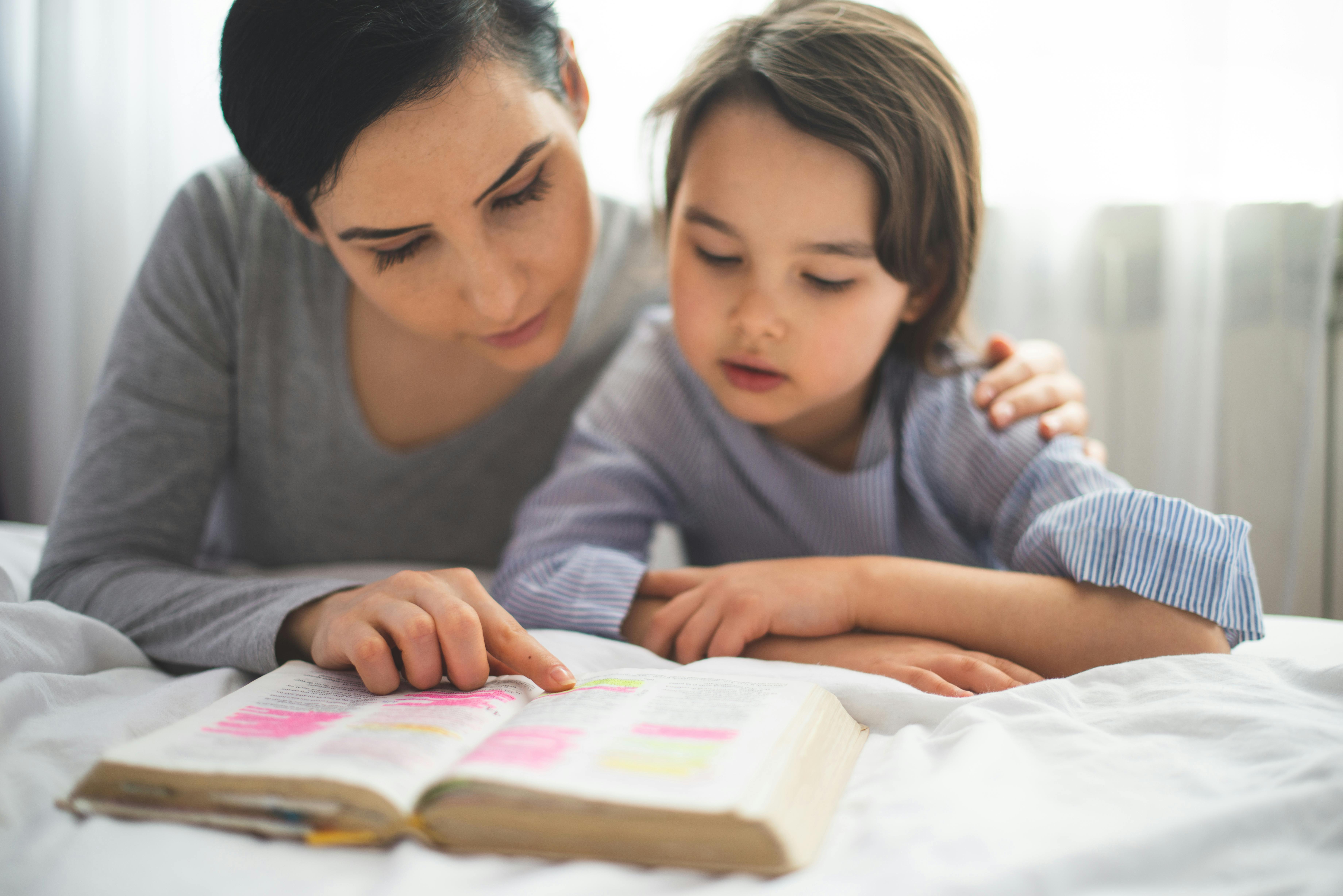 Une mère attentive et son jeune fils lisent ensemble un livre sur un lit, dans un moment de calme et d'apprentissage partagé. La mère pointe du doigt une page du livre, qui semble contenir des passages importants surlignés, tandis que l'enfant, concentré, suit attentivement. Ce moment capture une interaction éducative et affectueuse entre parent et enfant.