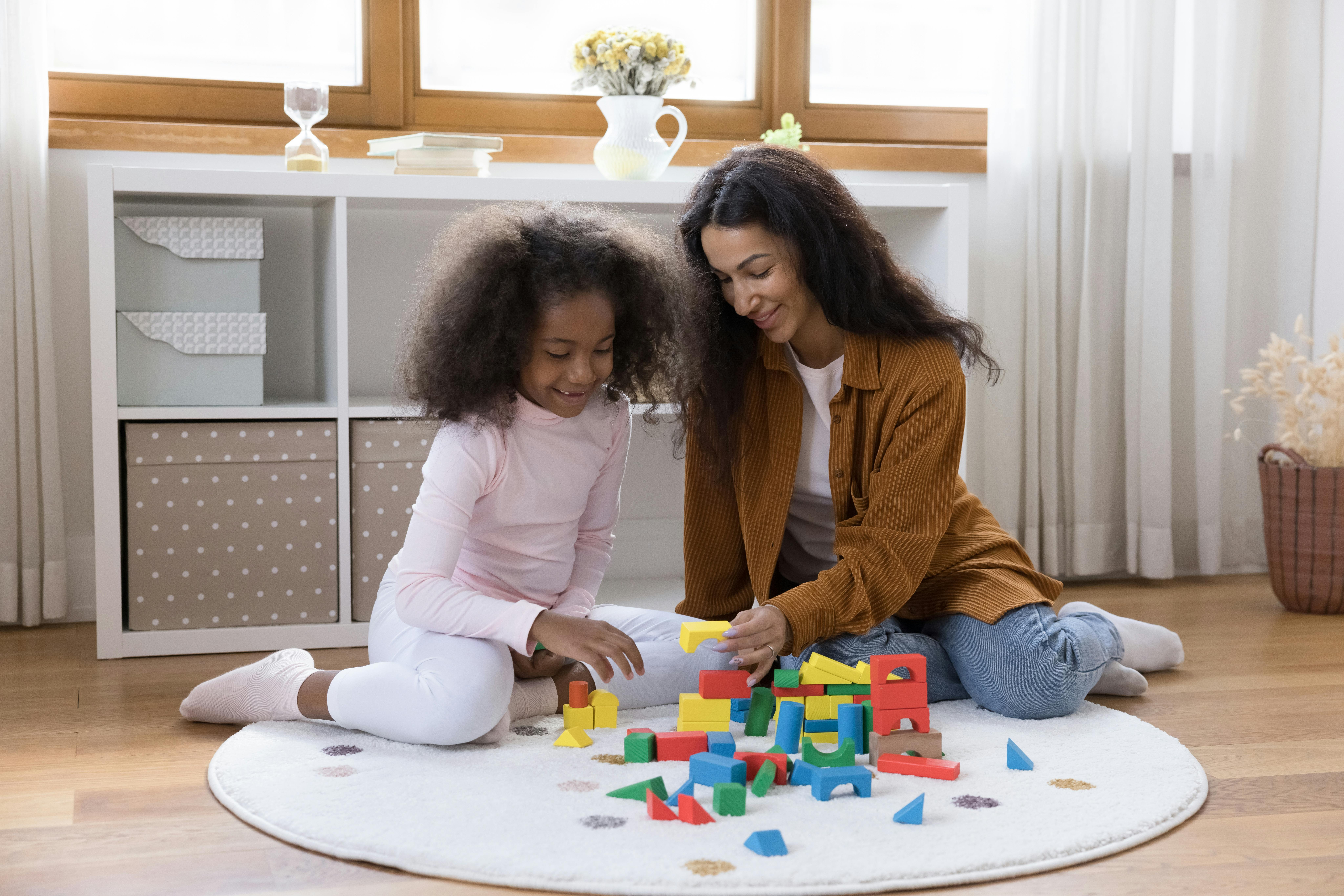 Une mère et sa fille jouent ensemble avec des blocs colorés sur un tapis circulaire blanc, souriantes et concentrées sur leur activité.