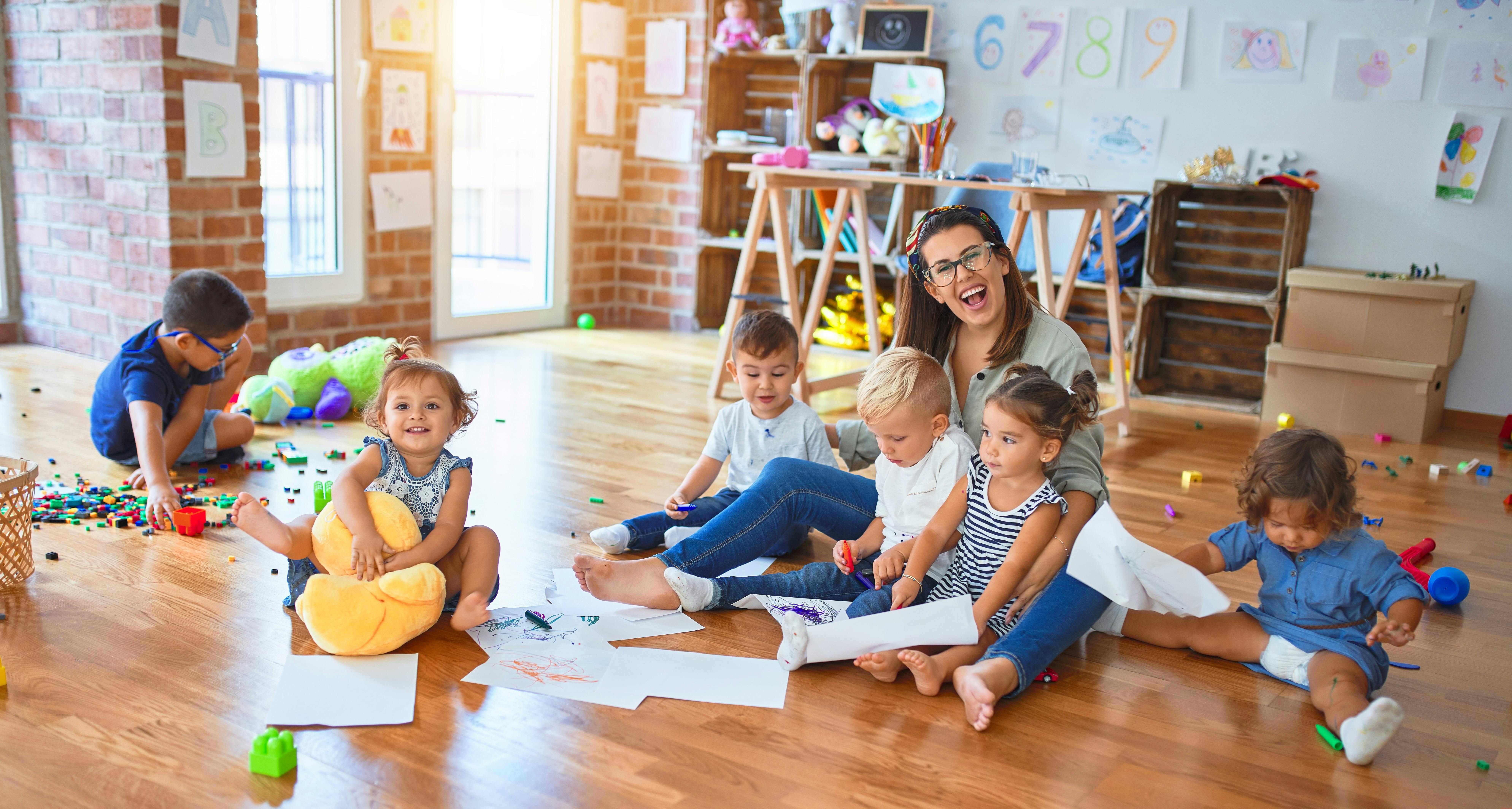 Une éducatrice entourée de jeunes enfants, participent à des activités créatives sur le sol d'une salle de jeux.