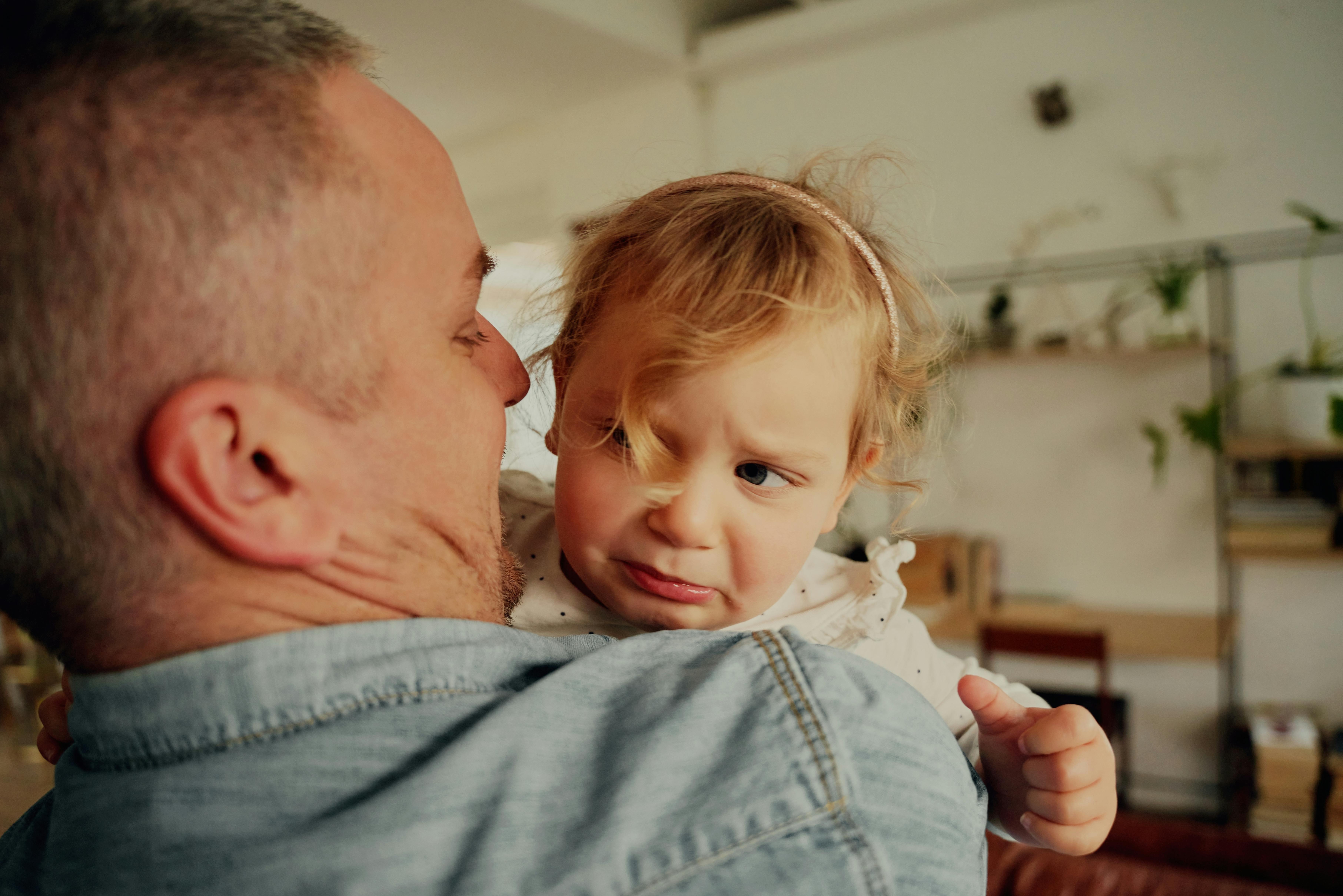 Un père tient dans ses bras une petite fille qui semble contrariée.